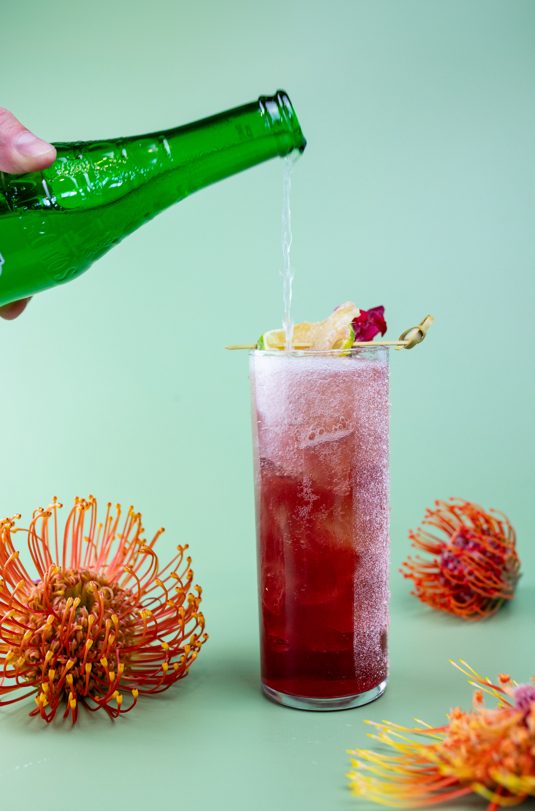 hibiscus ginger paloma with a bottle of jarritos grapefruit soda being poured into the glass, with a variety of flowers surrounding the cocktail.